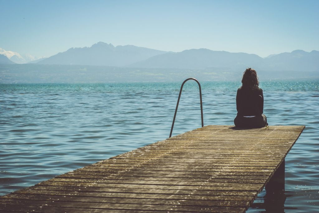 Photo of a desolate scene of a woman sitting on the edge of a dock looking out across a lake